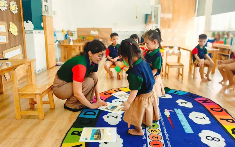 A teacher kneels on an alphabet mat to look at a picture card with two toddlers